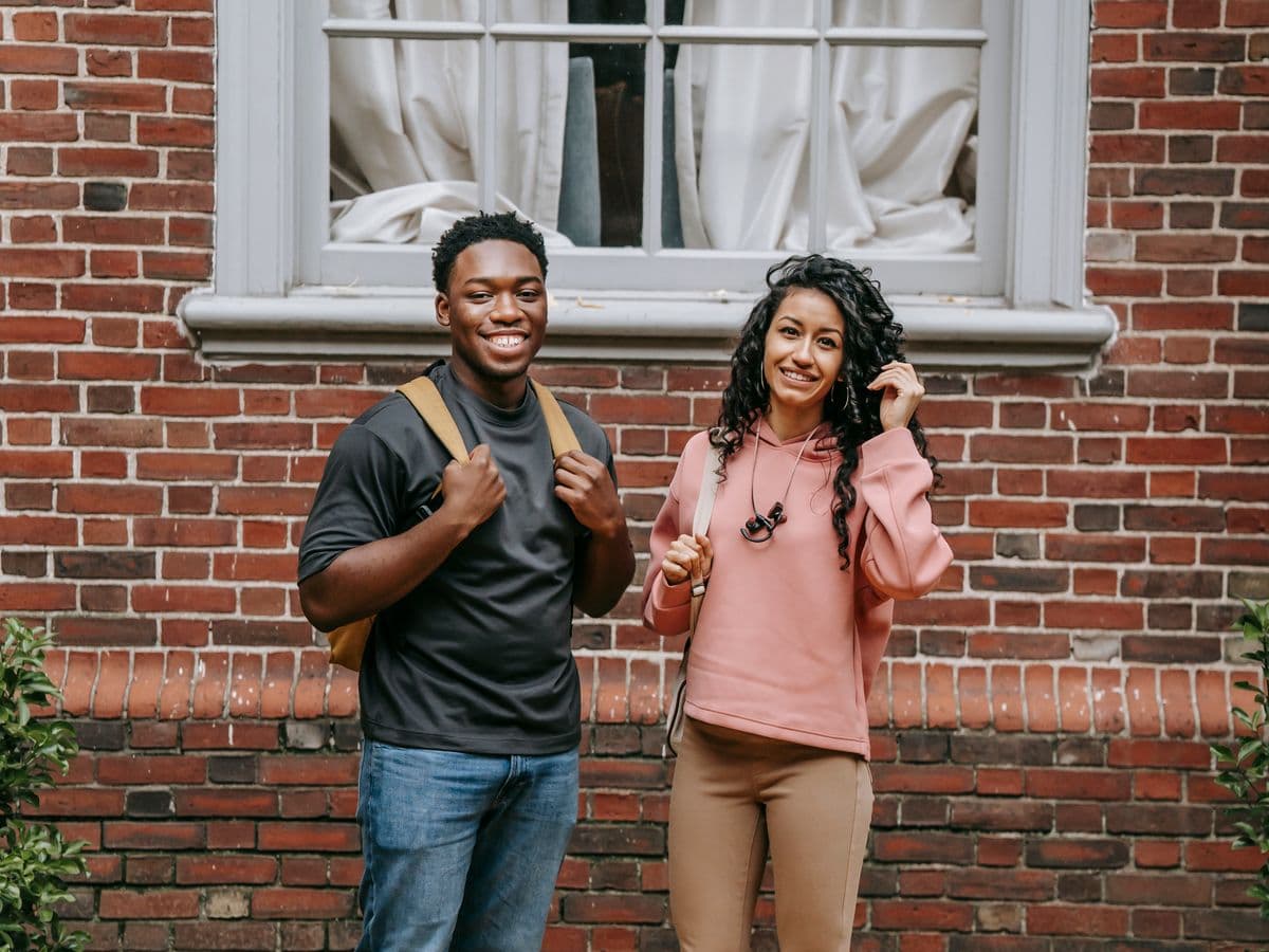 Deux étudiants souriants avec leurs sacs devant un bâtiment de campus
